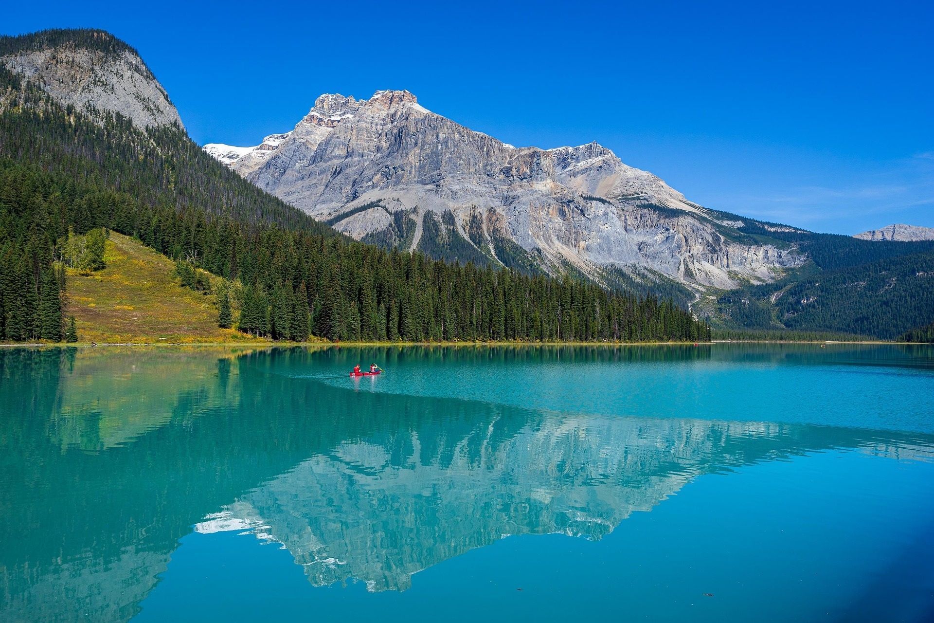 Der türkisfarbene See spiegelt schneebedeckte Berge und grüne Bäume unter einem strahlend blauen Himmel wider.