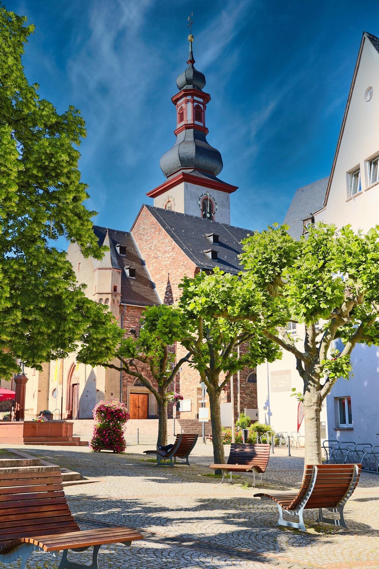 In Rüdesheim, auf einem Stadtplatz mit grünen Bäumen und Holzbänken unter blauem Himmel, steht eine Kirche mit einem verzierten Turm.