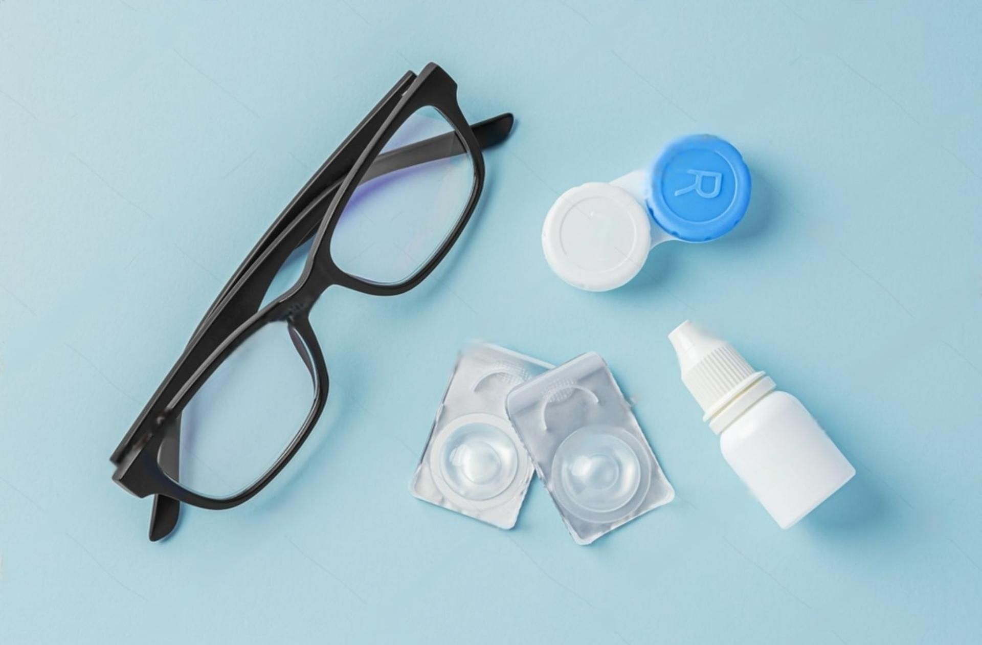Schwarze Brille, Kontaktlinsen, Lösung und Etui auf blauem Untergrund.