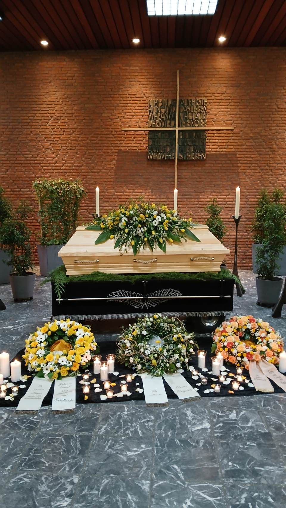 Ein heller Holzsarg, geschmückt mit Blumen, umgeben von Kränzen, Kerzen und Pflanzen in einer Trauerkulisse.