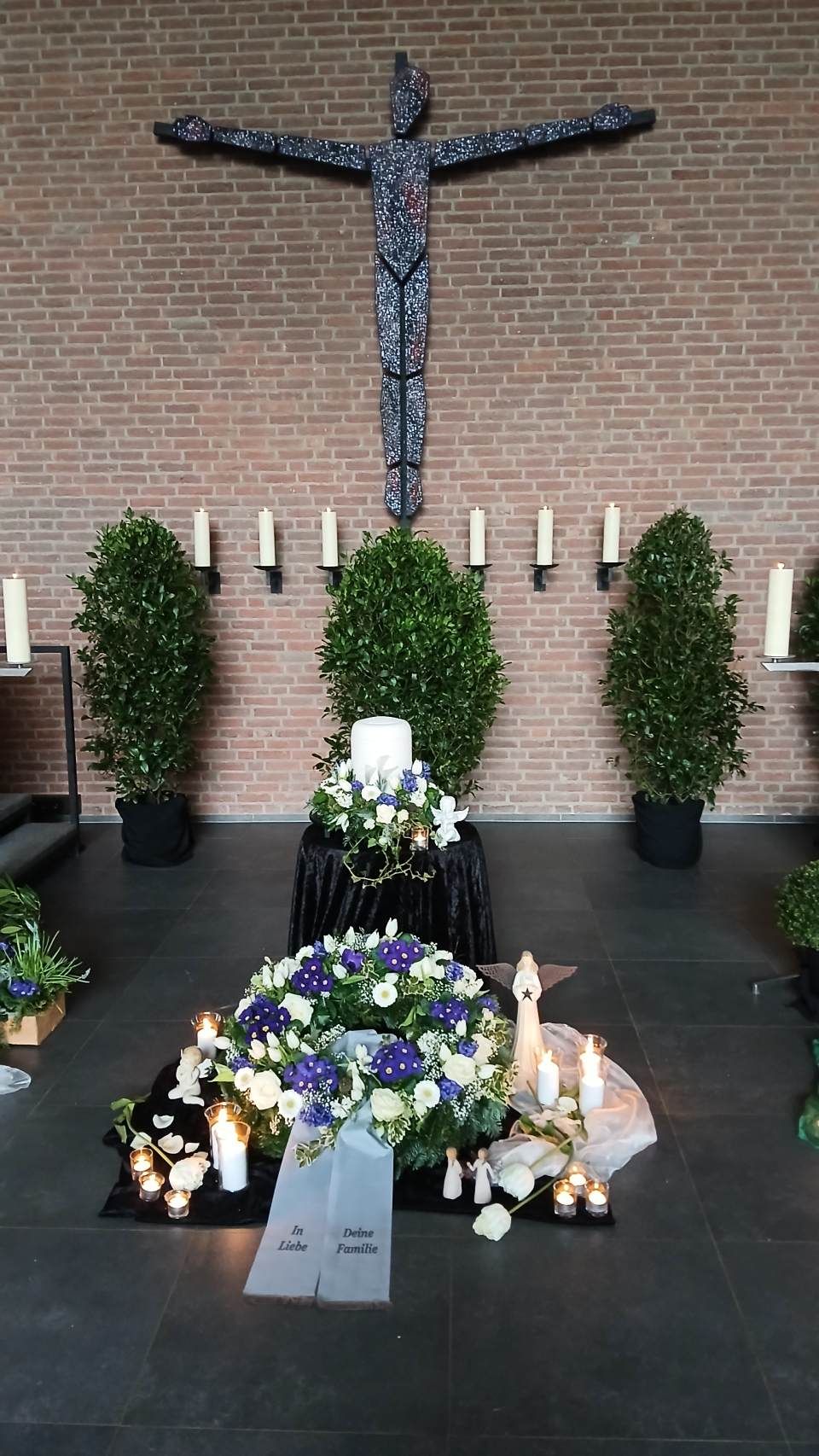Traueraltar mit Blumenkränzen, Kerzen und einer Kreuzskulptur. Backsteinmauer im Hintergrund.