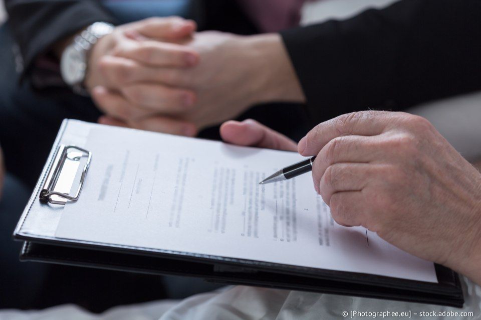 Eine Person hält einen Stift über ein Dokument auf einem Klemmbrett; im Hintergrund sind die Hände einer anderen Person zu sehen.