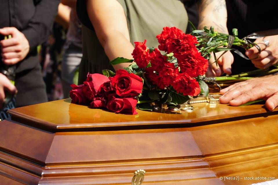 Rote Rosen liegen auf einem geschlossenen Holzsarg, daneben Hände und Arme von Menschen.