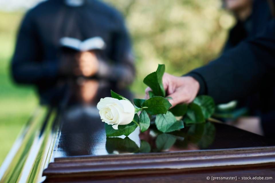 Eine Hand legt bei einer Beerdigung eine weiße Rose auf einen geschlossenen Sarg. Im Hintergrund steht eine Person in Schwarz.