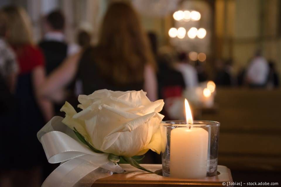 Weiße Rose und brennende Kerze in einem Glashalter bei einer Gedenkfeier. Menschen im Hintergrund.