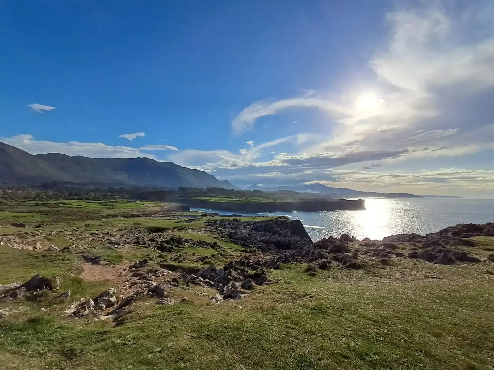 Paisaje costero con primer plano cubierto de hierba, costa rocosa, montañas y un sol brillante