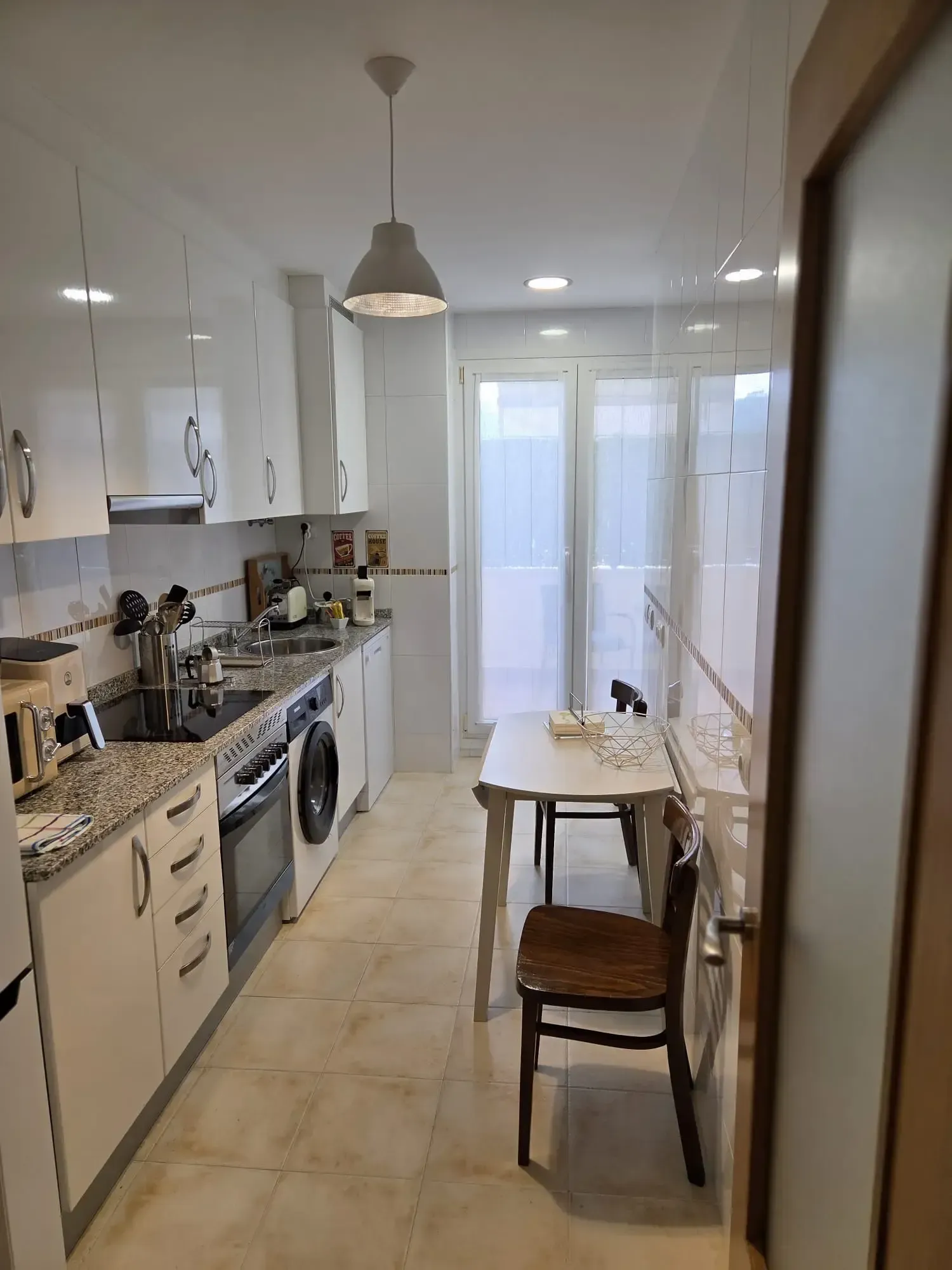 Cocina estrecha de color blanco con suelo de baldosas, fogones, fregadero, mesa pequeña y silla cerca de una puerta luminosa.