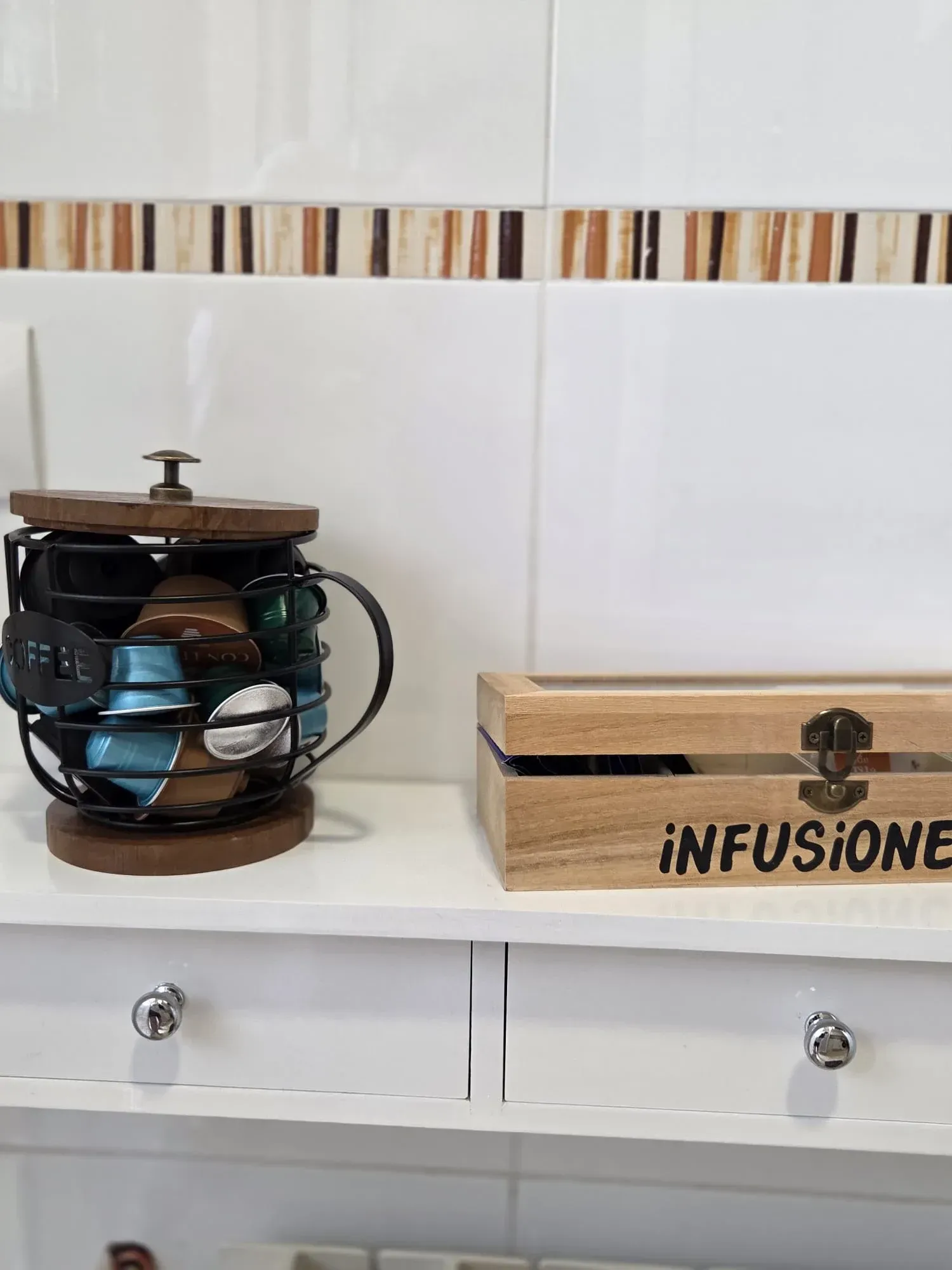 Encimera con soporte para cápsulas de café y caja infusora de madera sobre un estante de cocina alicatado.