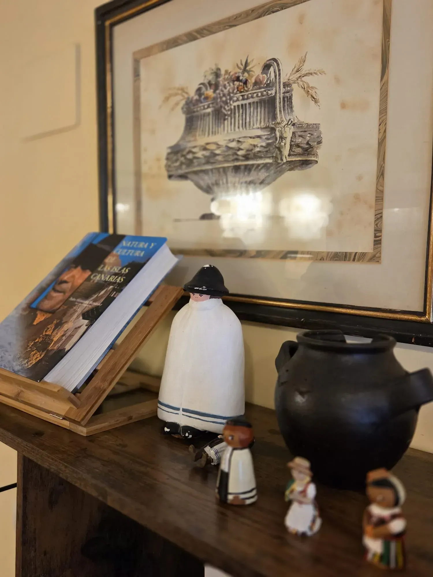 Libros y figuritas pequeñas sobre un estante de madera debajo de un boceto enmarcado y un jarrón negro.