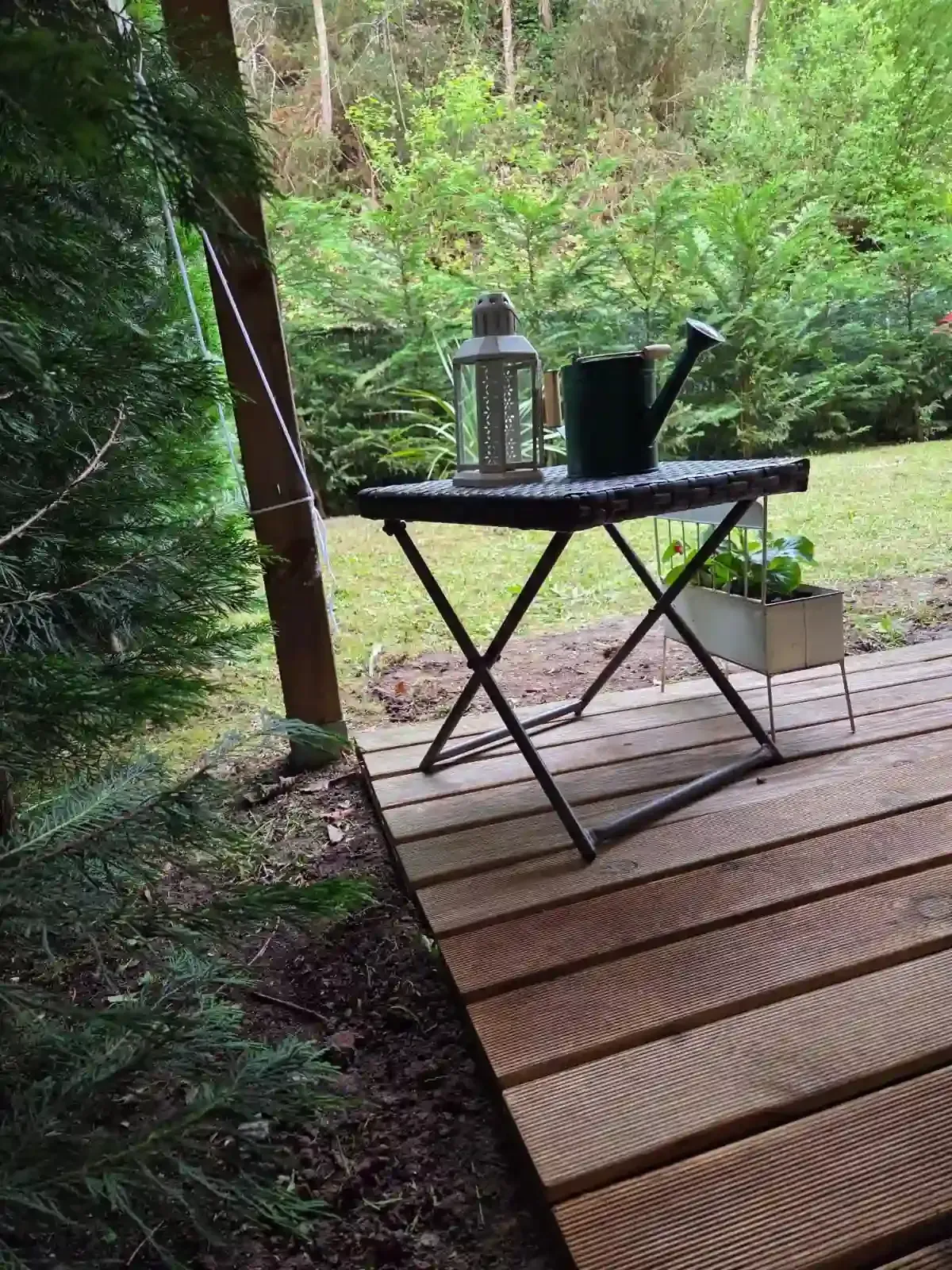 Mesita sobre una terraza de madera en un bosque frondoso, con una linterna y una regadera junto a un sendero.