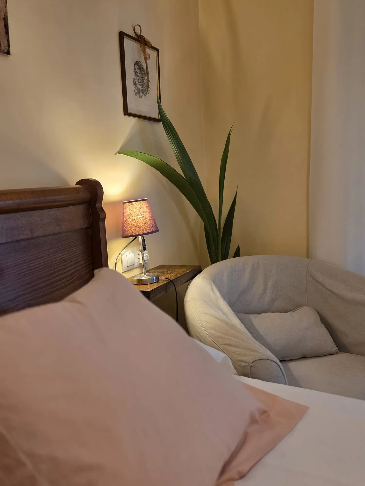 Acogedor rincón de dormitorio con cama, lámpara encendida, silla blanca y planta alta contra una pared beige.
