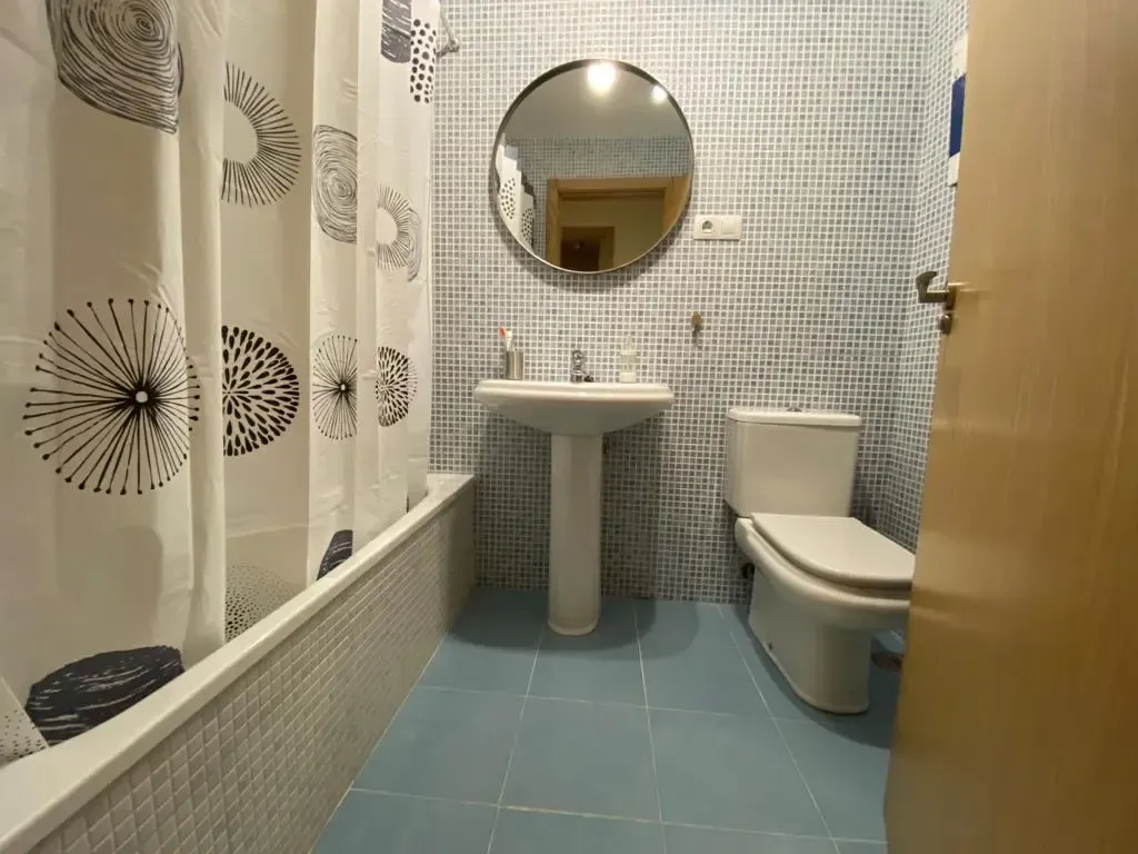 Baño pequeño con azulejos, lavabo, inodoro, cortina de ducha, espejo y baldosas azules en el suelo.