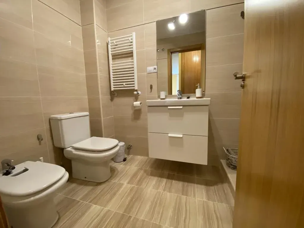 Baño moderno con azulejos beige, inodoro, bidé, lavabo suspendido, espejo y radiador toallero.