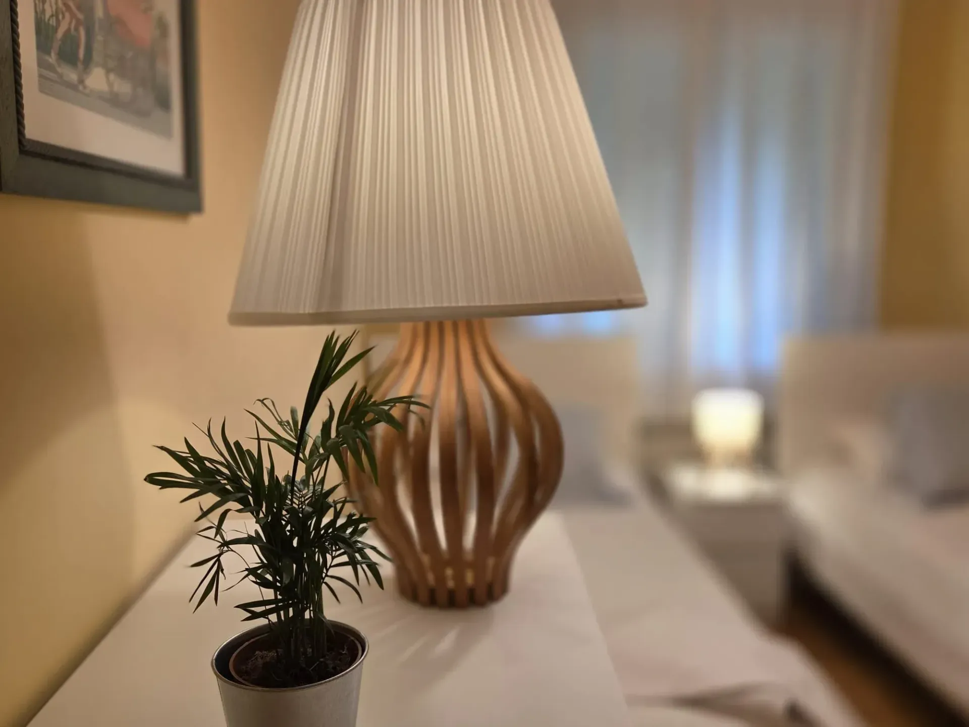 Lámpara de mesa y una pequeña planta en maceta sobre una mesita auxiliar blanca en una sala de estar con iluminación tenue.