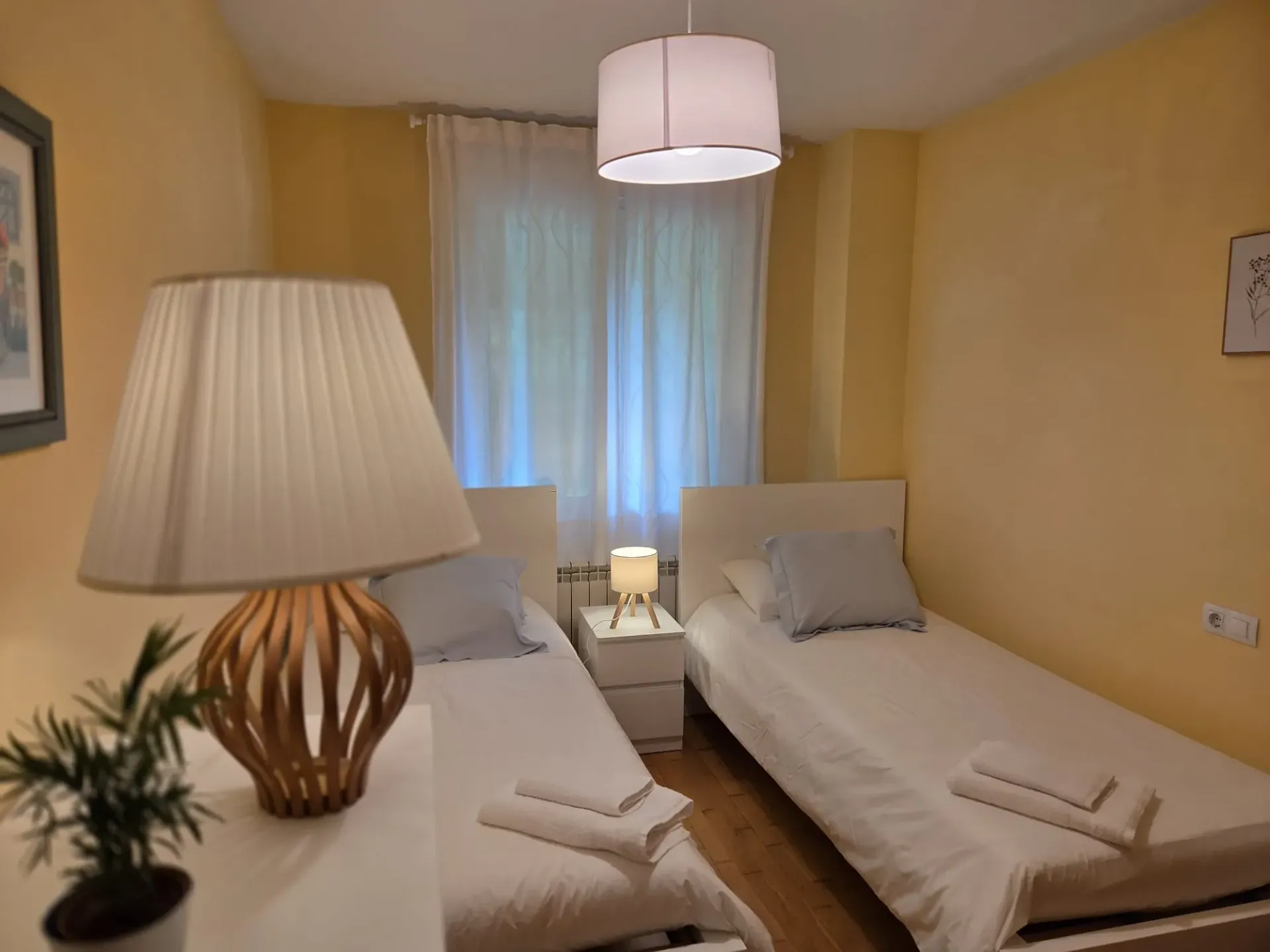 Habitación de hotel pequeña con dos camas individuales, paredes amarillas, una lámpara de noche y una ventana con cortinas.