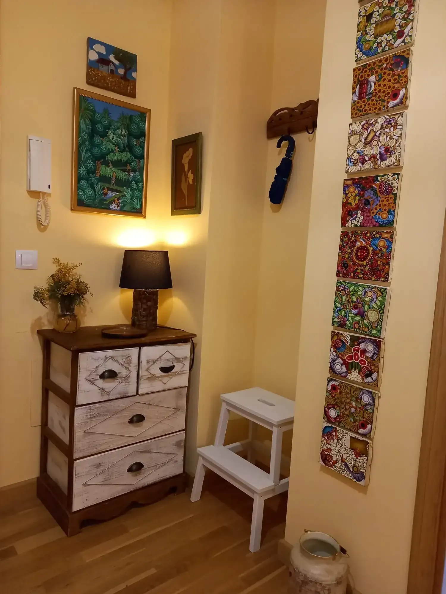 Rincón acogedor con cómoda de madera, lámpara encendida, taburete pequeño, cuadro en la pared y tapiz colorido colgante.