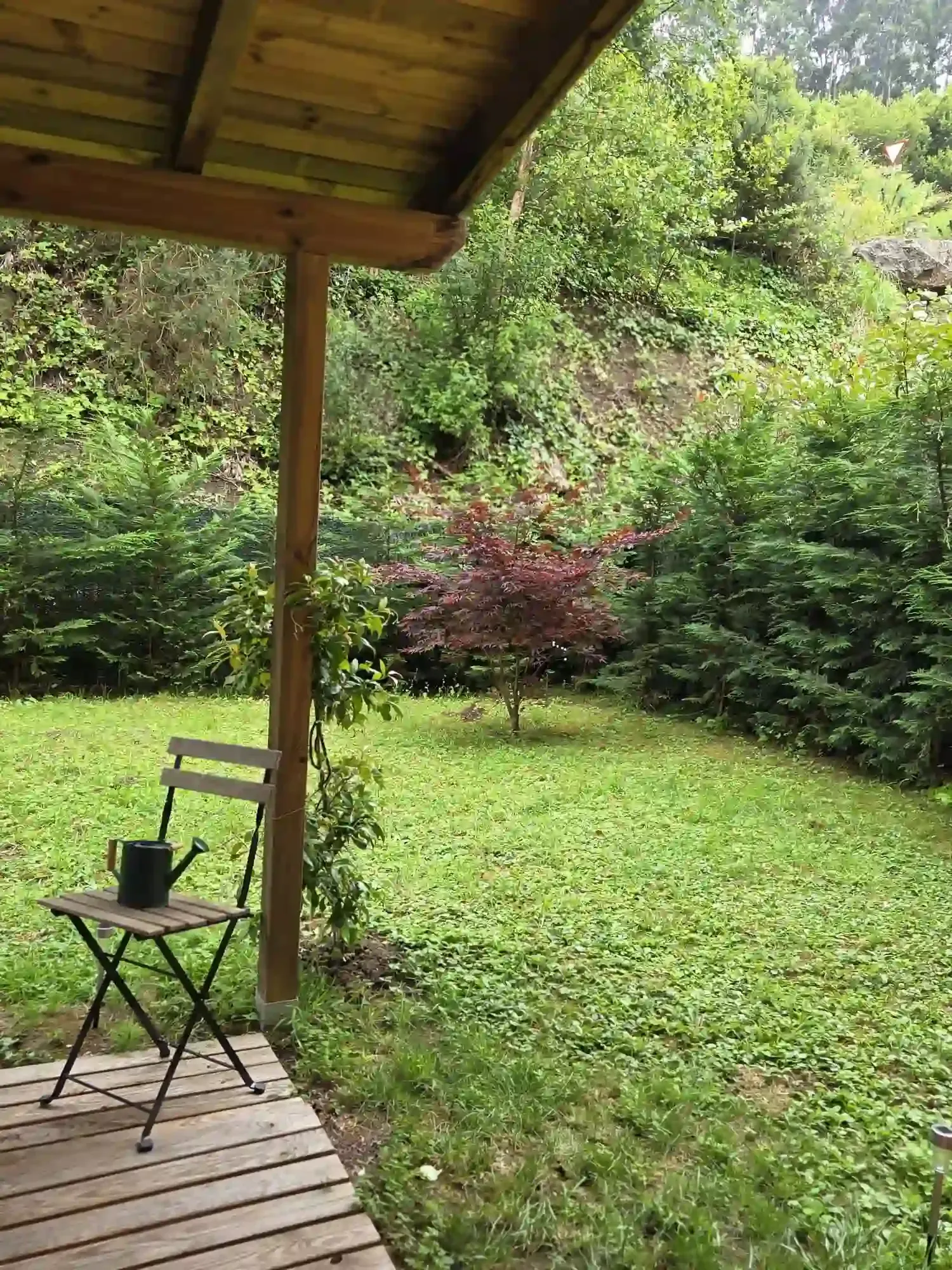 Patio de madera cubierto con vistas a un jardín verde con arbustos y una pequeña mesa y silla negras.