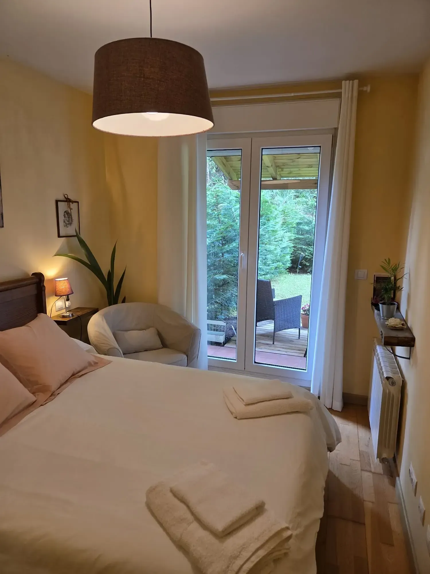 Acogedora habitación con cama blanca, lámpara marrón, silla junto a las puertas de cristal y vegetación en el exterior.