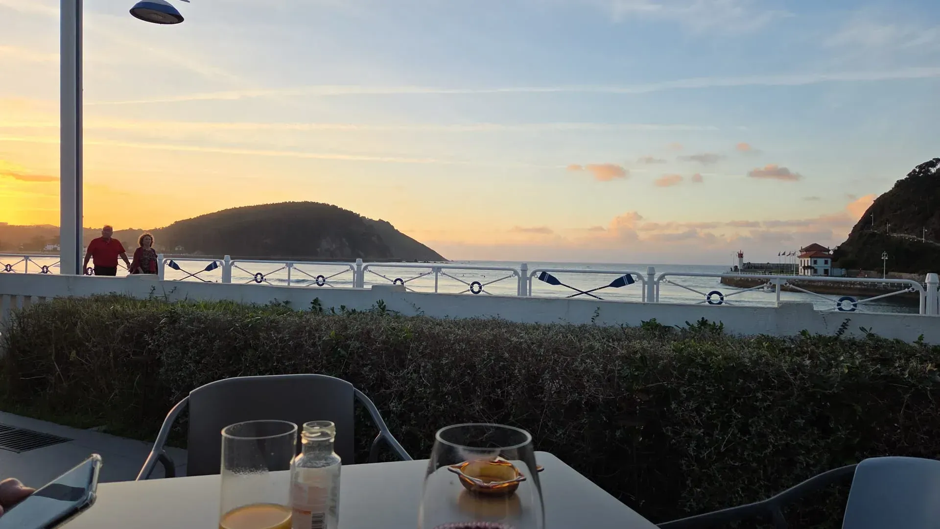 Vista del atardecer desde una cafetería junto al mar con mesas, bebidas, un seto y, a lo lejos, barcos y colinas.