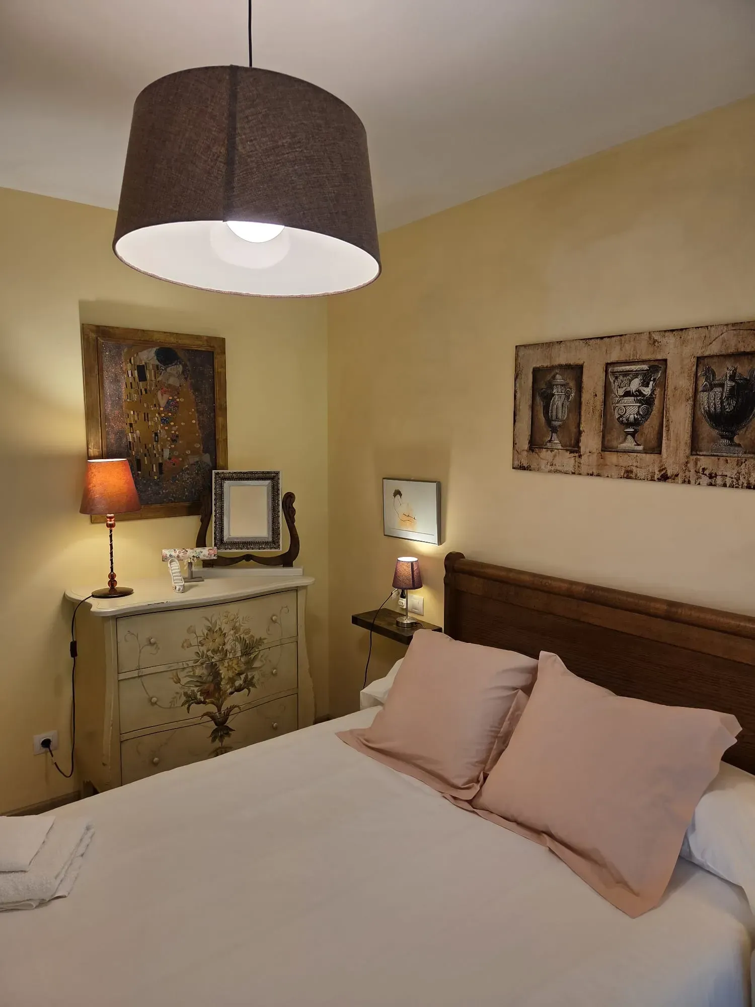 Acogedora habitación con cama blanca, paredes beige, lámparas marrones y un cuadro decorativo sobre una cómoda.
