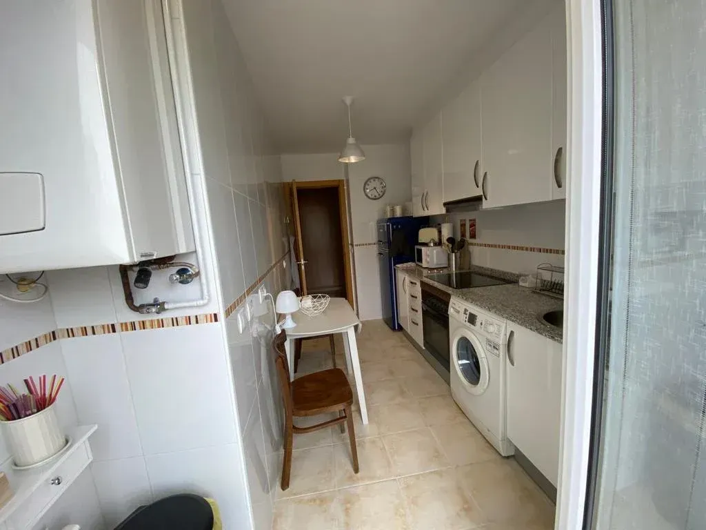 Cocina estrecha y blanca tipo galera con armarios, estufa, lavadora, fregadero y una mesa y silla pequeñas.