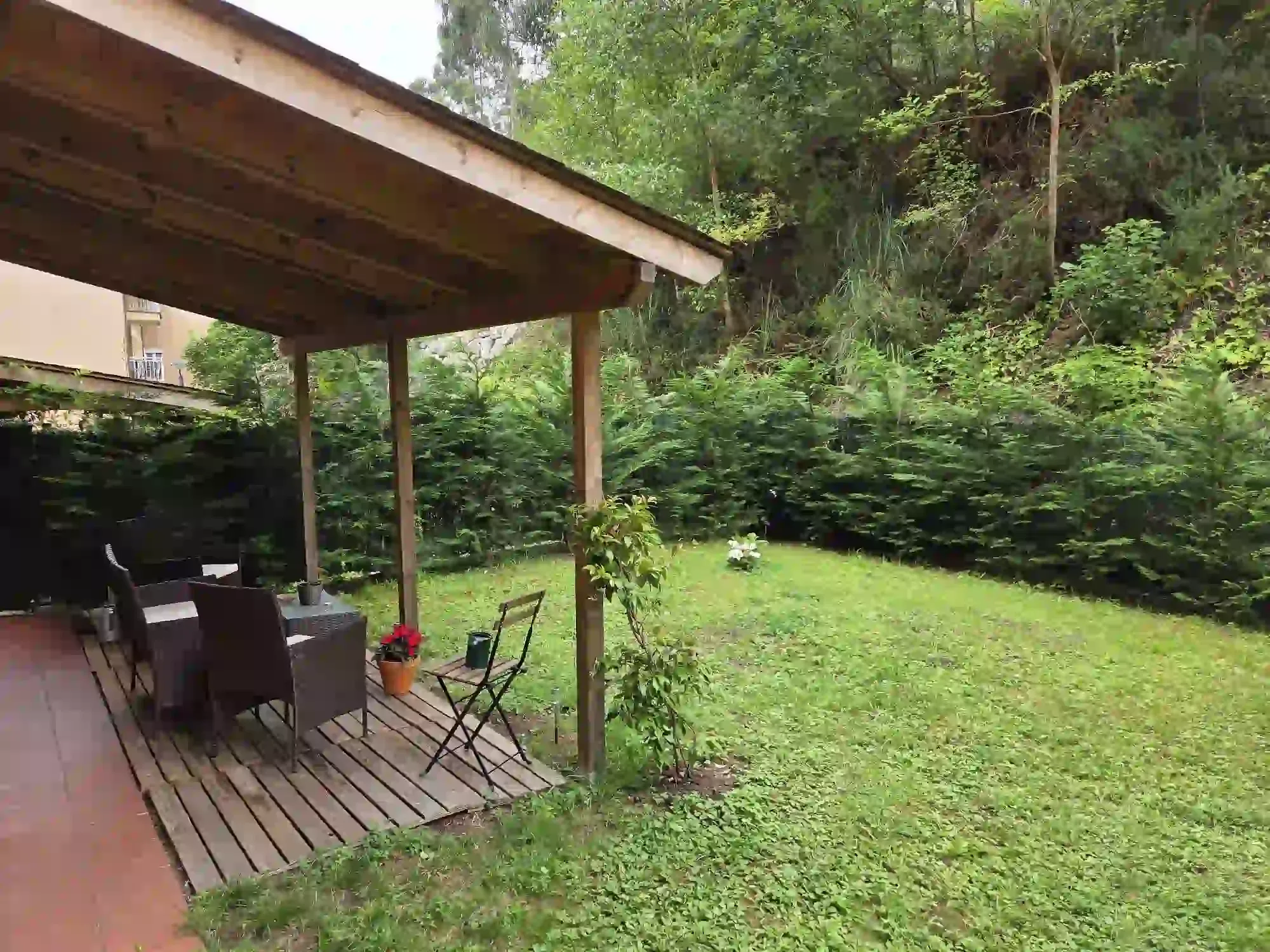 Patio cubierto con comedor al aire libre junto a un césped verde y un jardín arbolado.