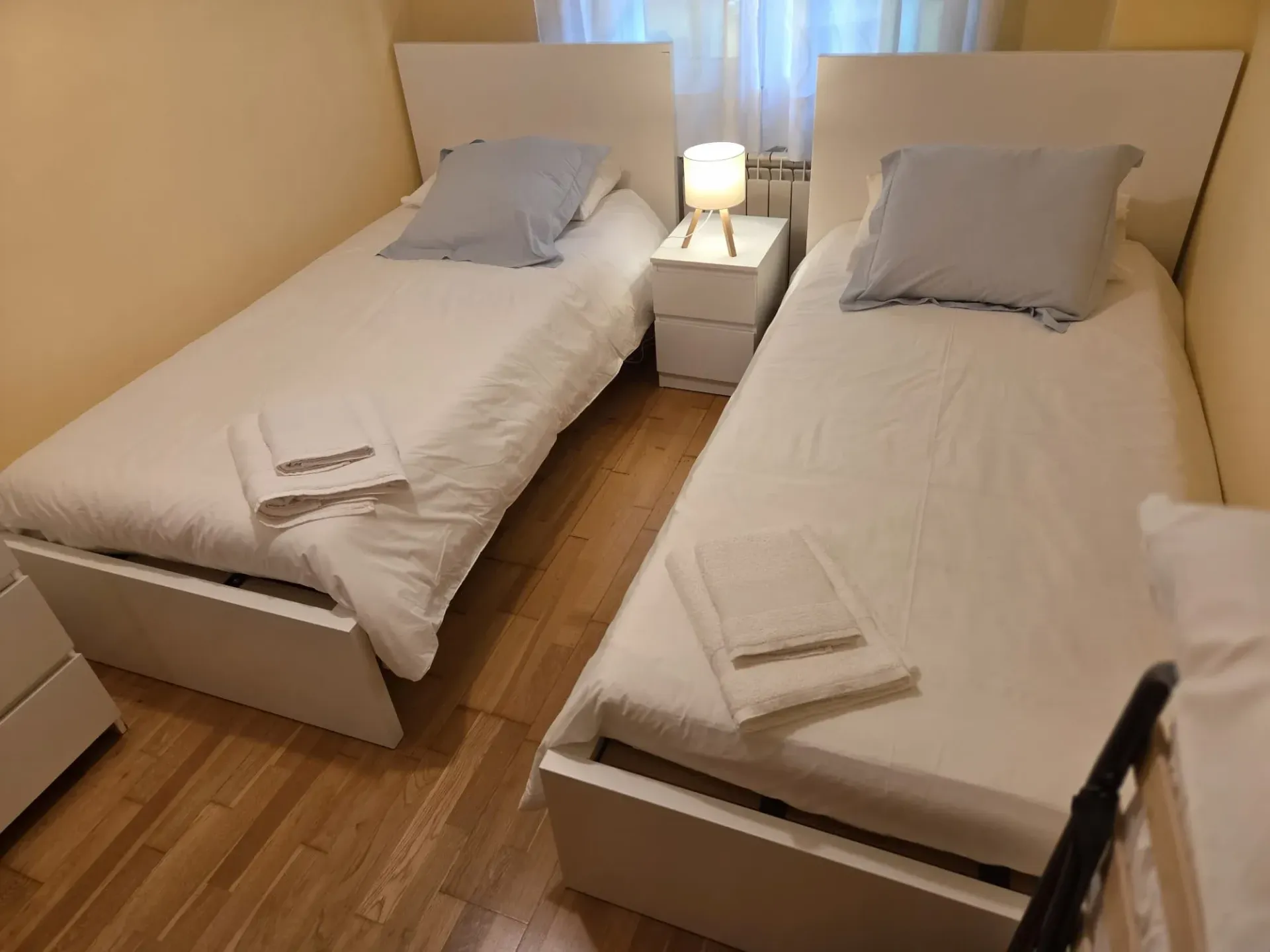 Dos camas individuales con ropa de cama gris en una habitación pequeña y ordenada con una lámpara de noche entre ellas.