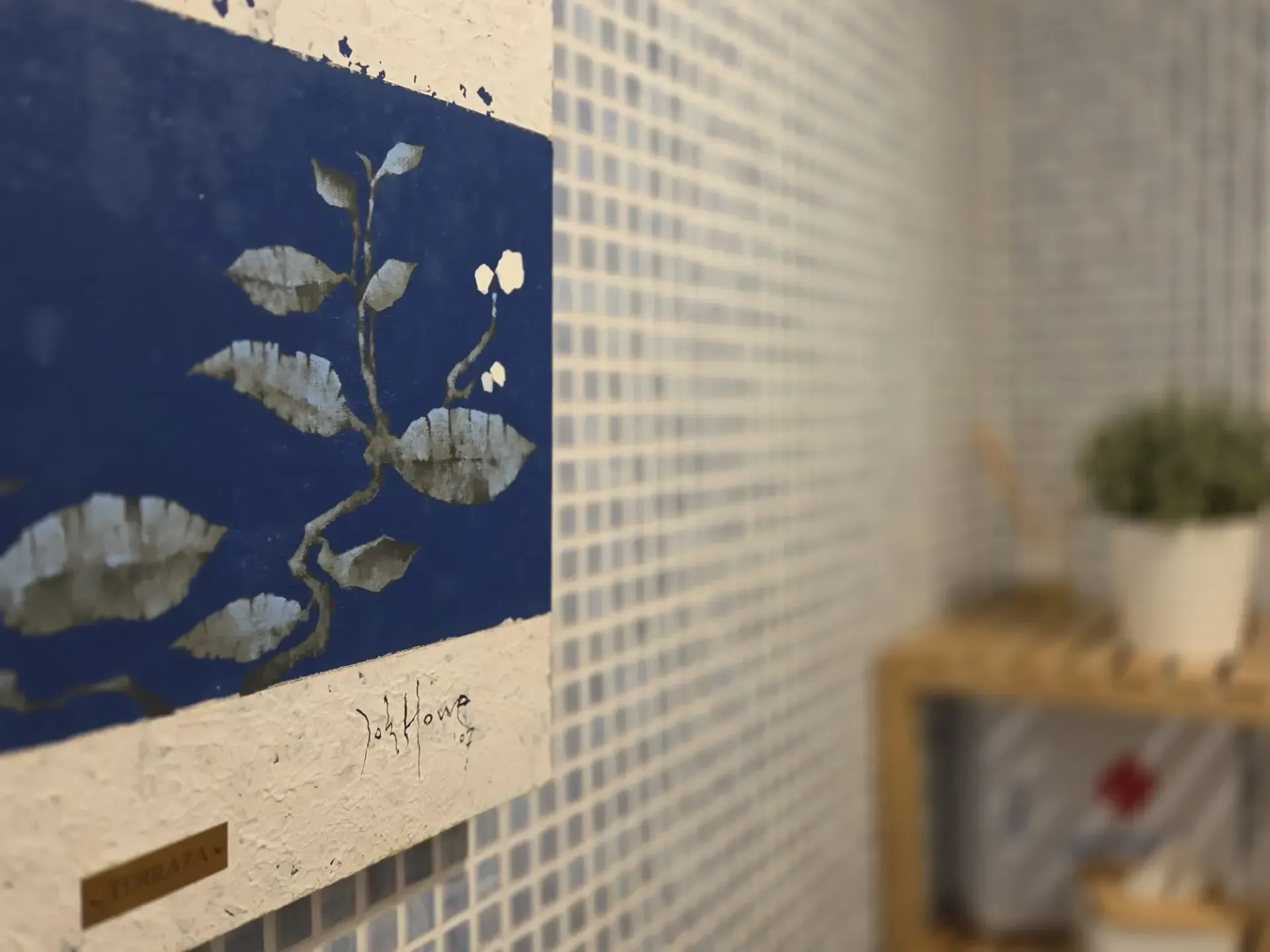 Cuadro azul con estampados de hojas blancas junto a una pequeña repisa con una planta y elementos decorativos.