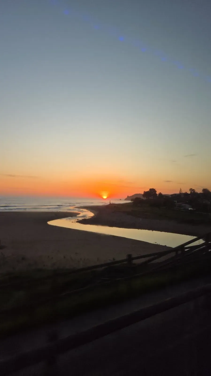 Puesta de sol sobre una playa con cielo anaranjado, aguas tranquilas y siluetas oscuras de la costa.