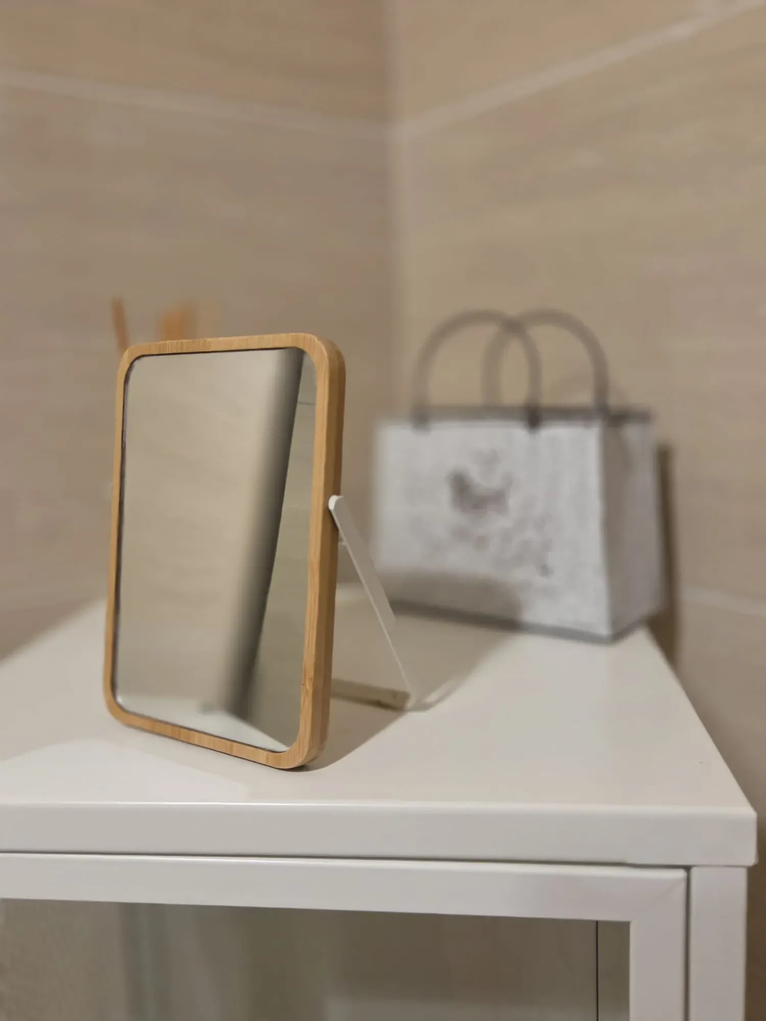 Un pequeño espejo de madera sobre una mesa blanca, con un bolso blanco difuminado al fondo.