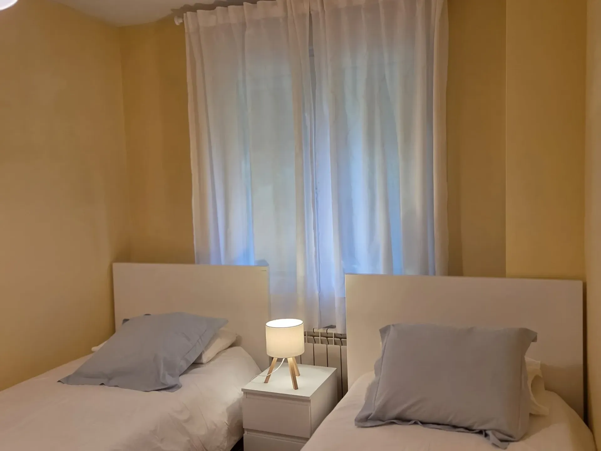Habitación pequeña con dos camas individuales, almohadas grises y una lámpara entre ellas, junto a la ventana.
