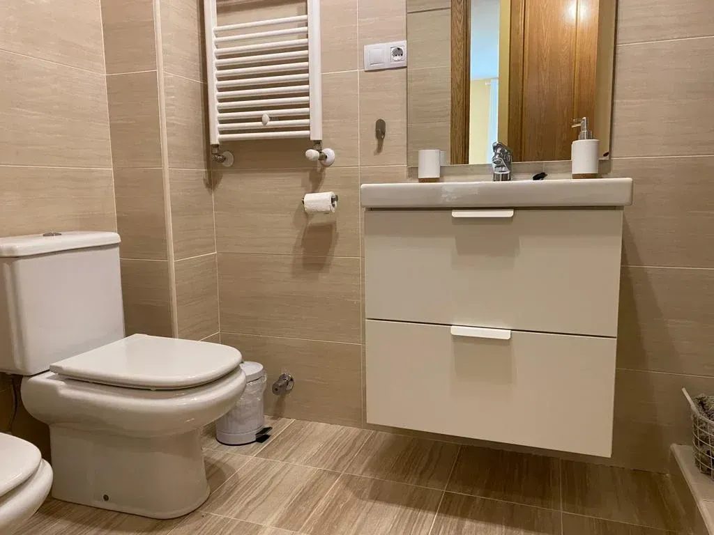 Baño moderno de color beige con inodoro, lavabo con mueble, espejo y toallero eléctrico.