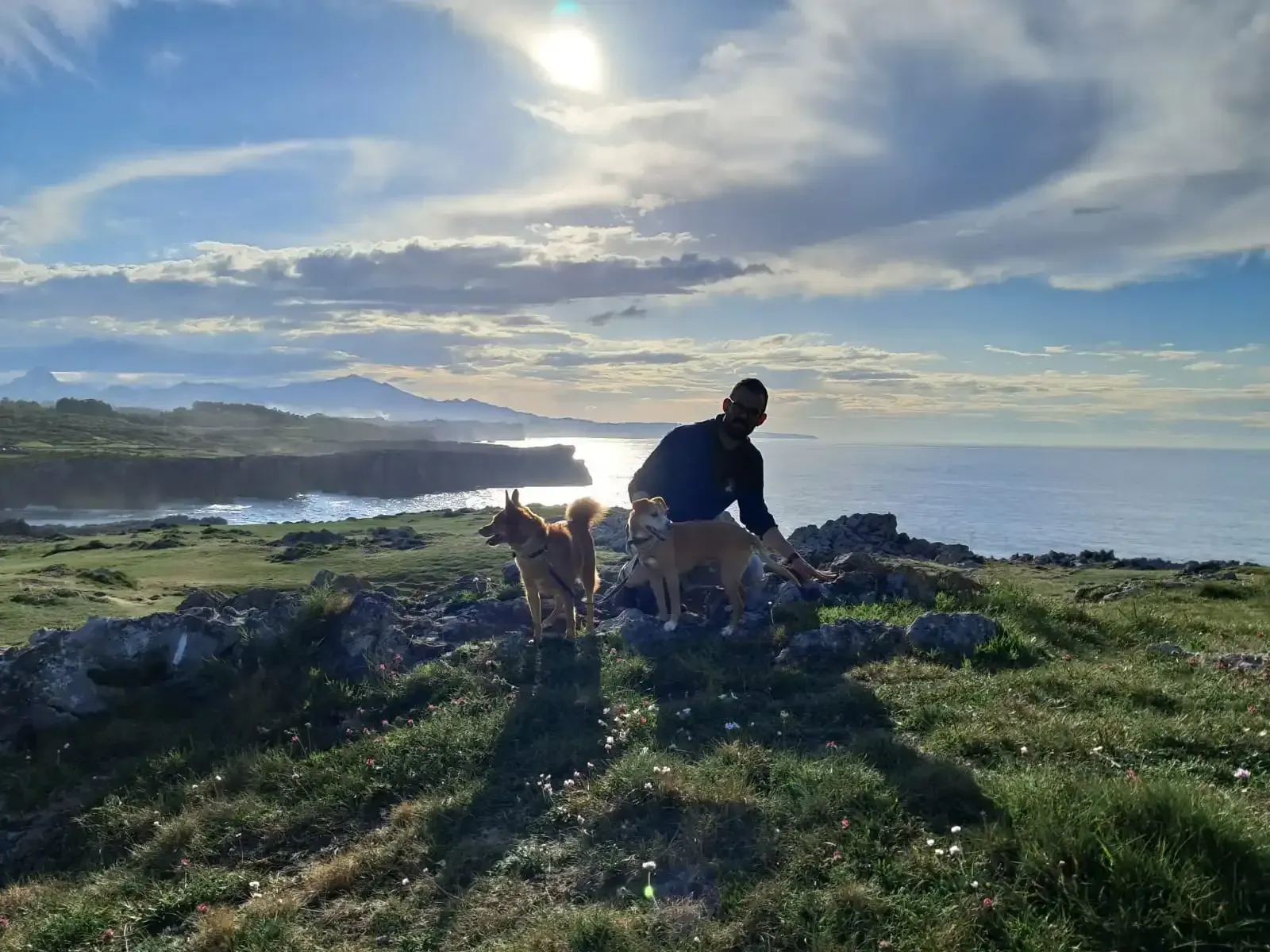 Una persona y su perro en un acantilado rocoso costero al atardecer, con el océano y un cielo brillante de fondo.