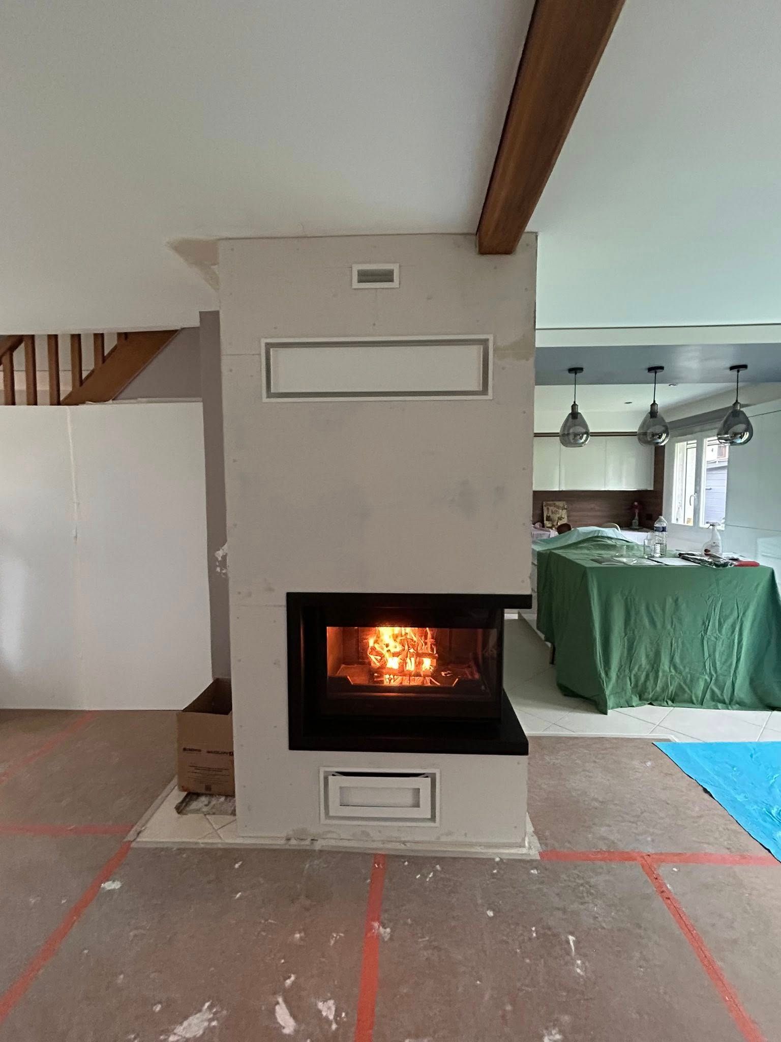 Cheminée moderne dans un salon, où crépite un feu éclatant, centrée sur un mur blanc aux poutres apparentes.