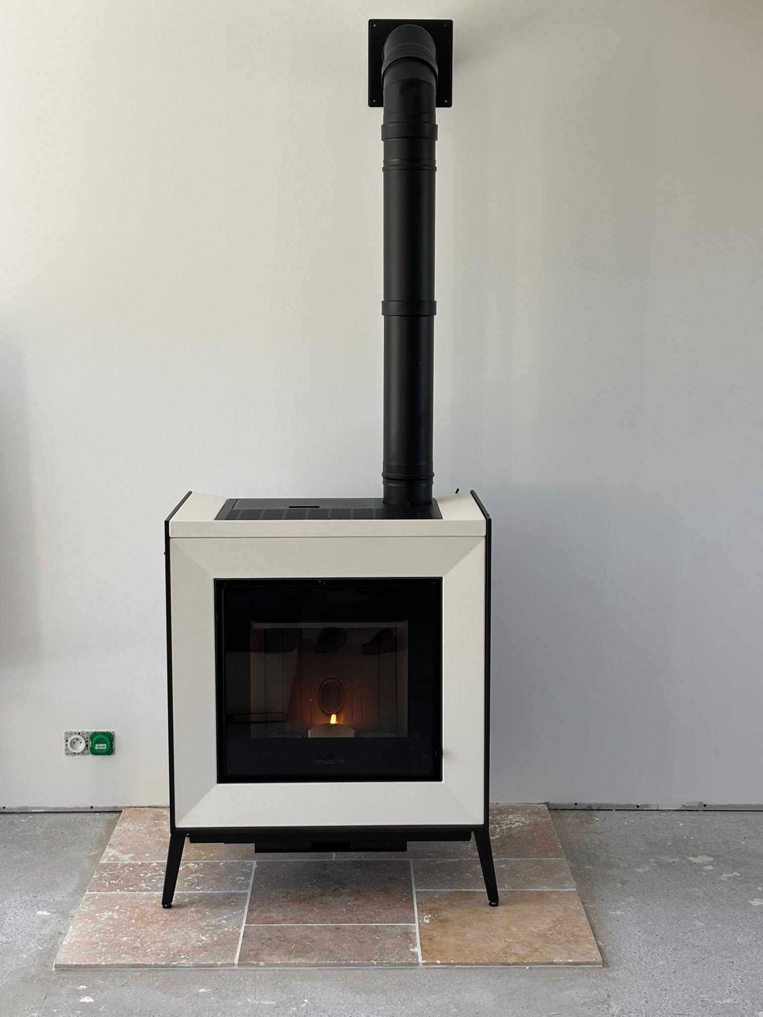 Poêle à bois blanc moderne avec conduit de cheminée contre un mur nu, sur un foyer carrelé.