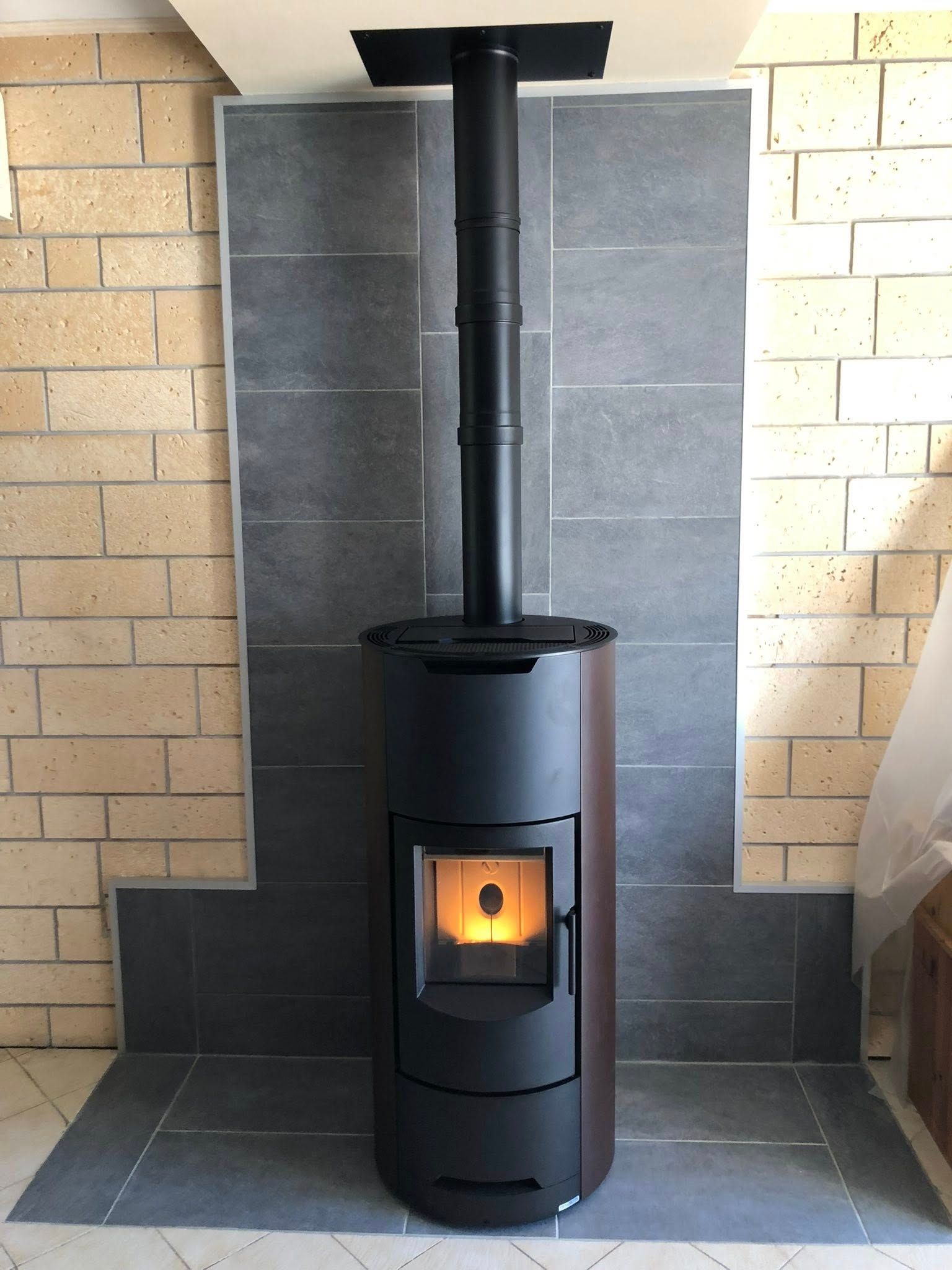 Poêle à granulés noir avec un feu incandescent, centré sur un foyer et un conduit de cheminée en pierre grise, contre un mur de briques.