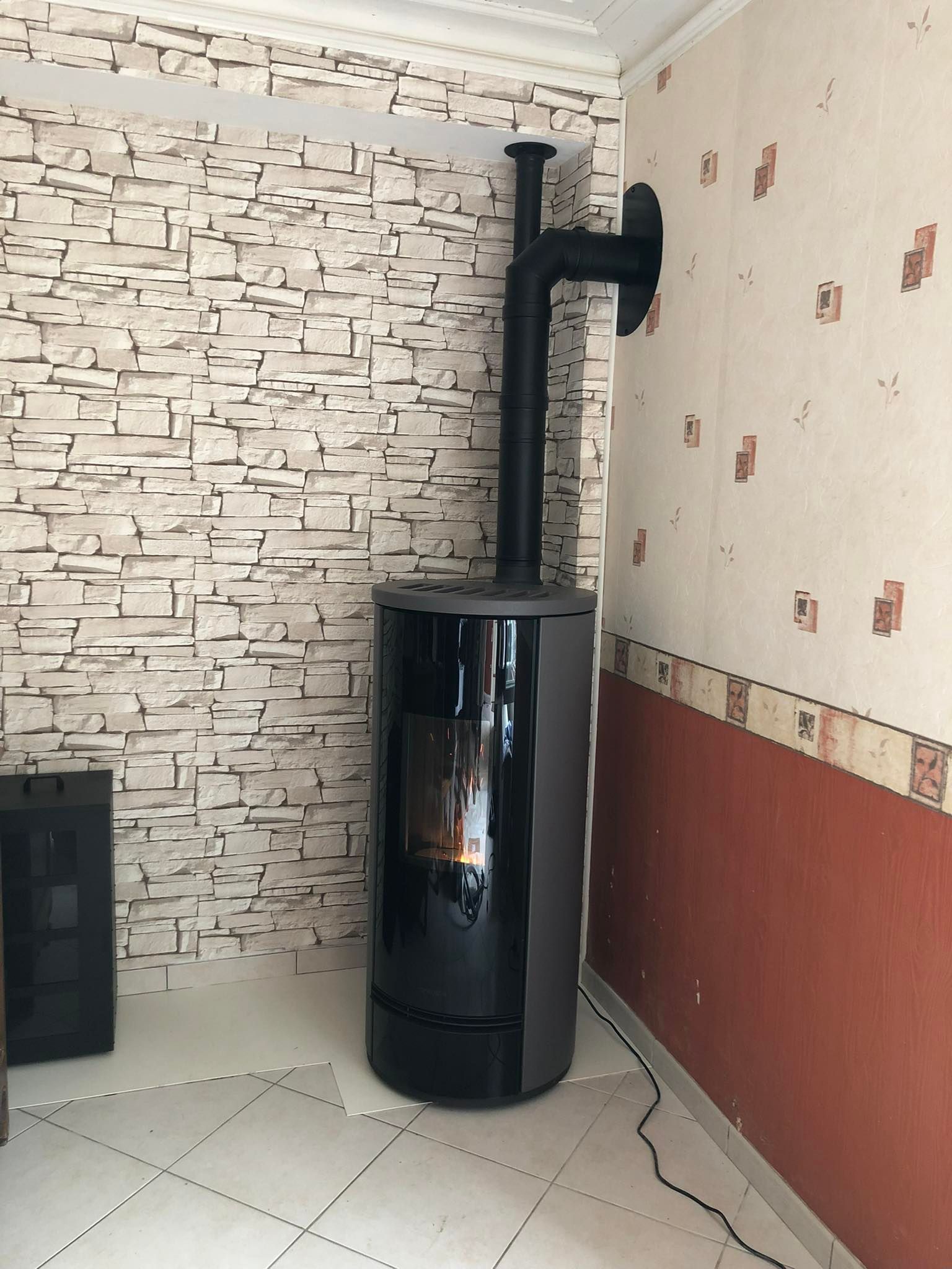 Poêle à bois noir avec porte vitrée et conduit de cheminée dans dans l'angle d'une pièce carrelée.