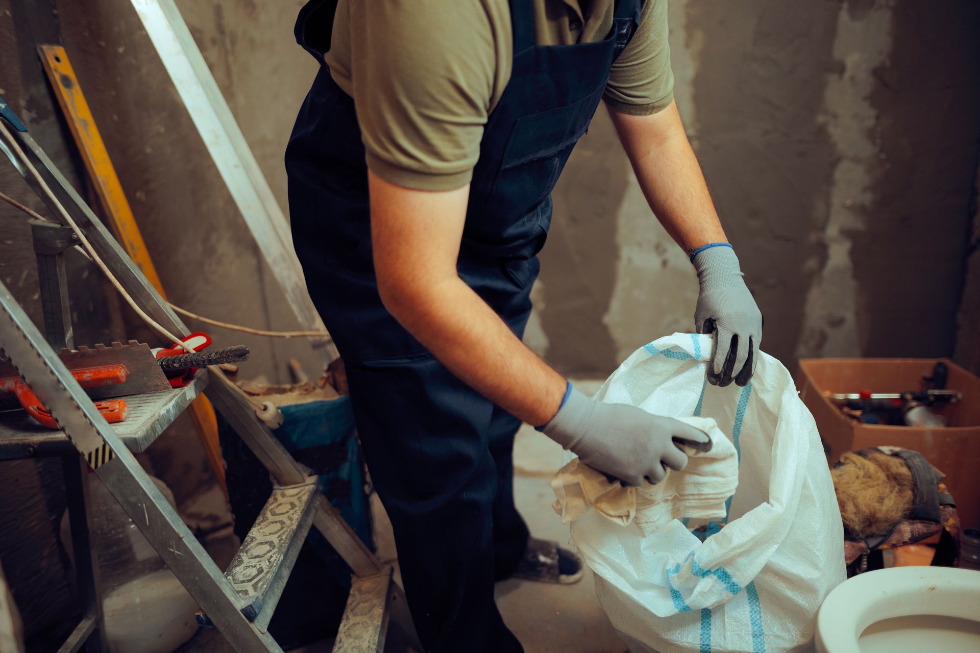 Ein Arbeiter mit Handschuhen öffnet einen weissen Sack in einem unordentlichen Baustellenbereich.