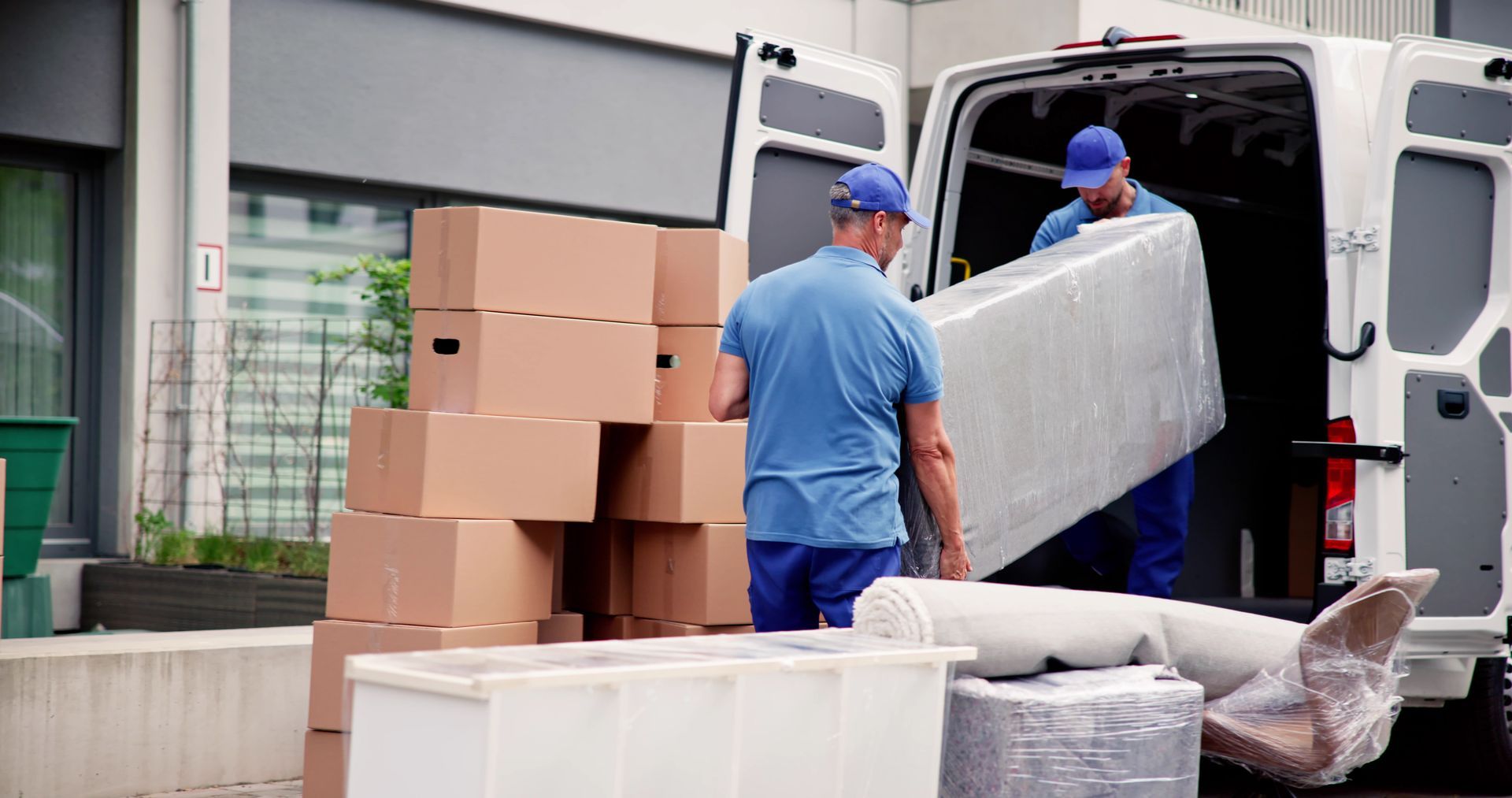Zwei Umzugshelfer verladen gestapelte Kartons und verpackte Möbel vor einem Gebäude in einen weissen Umzugswagen.