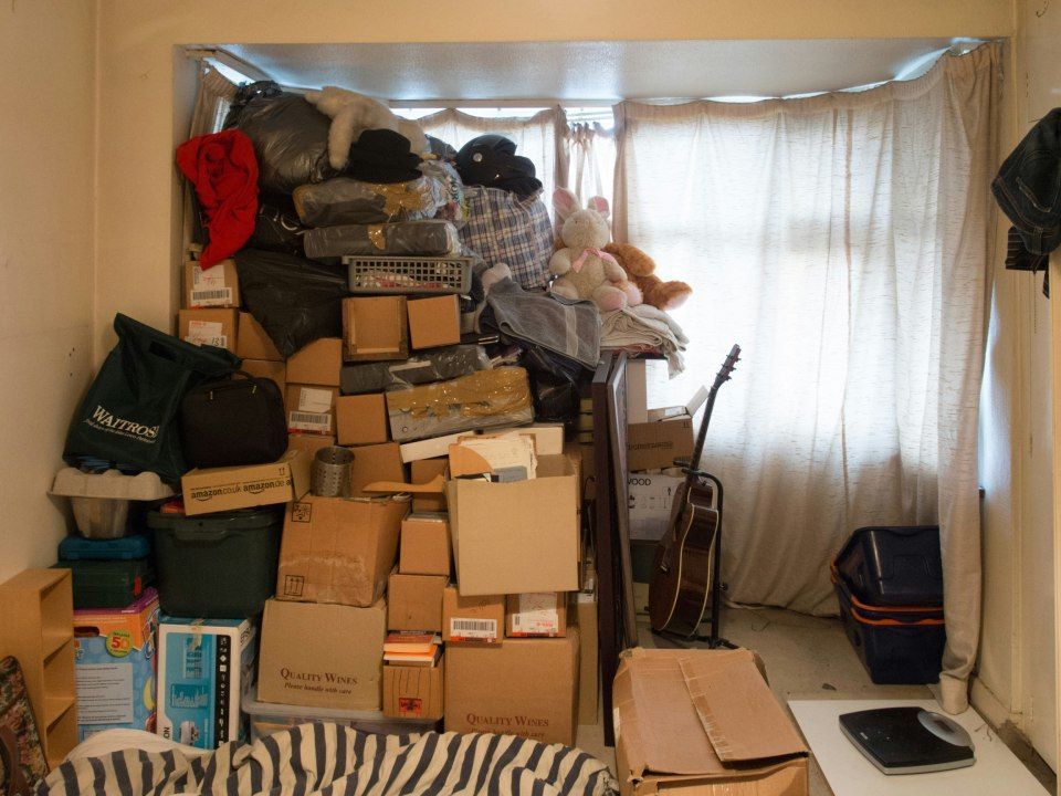 Ein unordentliches Zimmer mit hochgestapelten Pappkartons und Säcken neben einem Fenster mit Vorhängen.