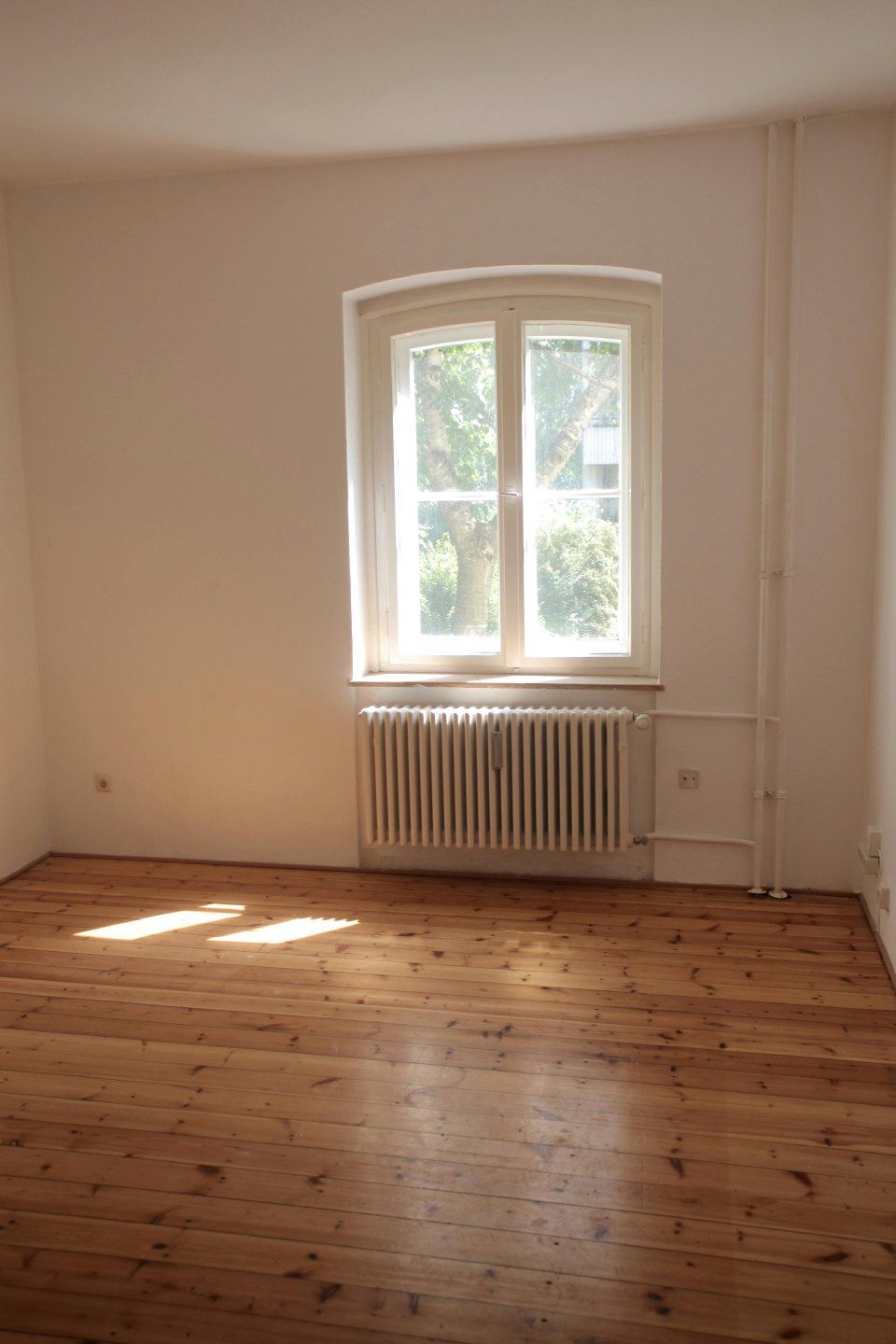 Leerer Raum mit Holzboden, weissen Wänden und einem Fenster über einem Heizkörper.