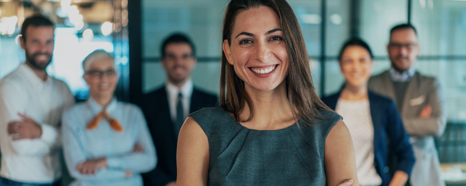 Une femme souriante en haut gris se tient dans un bureau, ses collègues étant flous en arrière-plan.