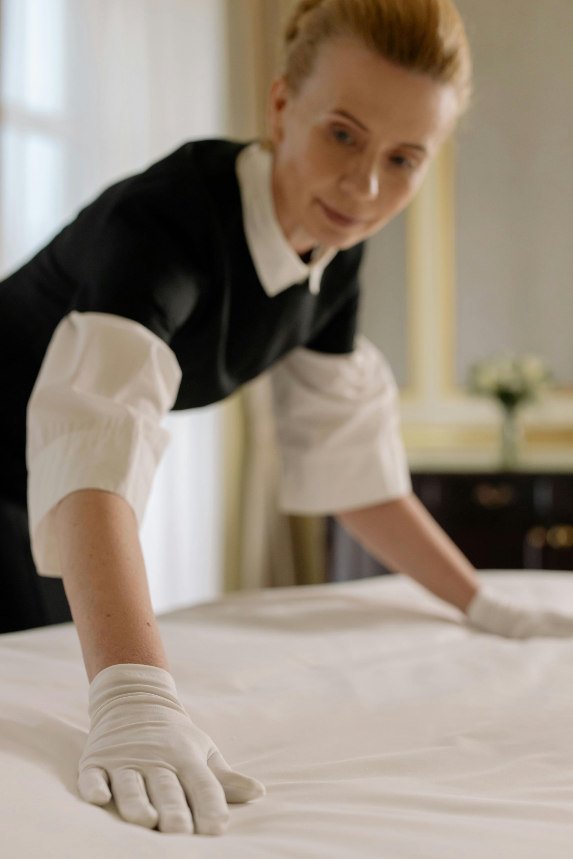 Femme de ménage en gants blancs faisant un lit dans une chambre d'hôtel lumineuse