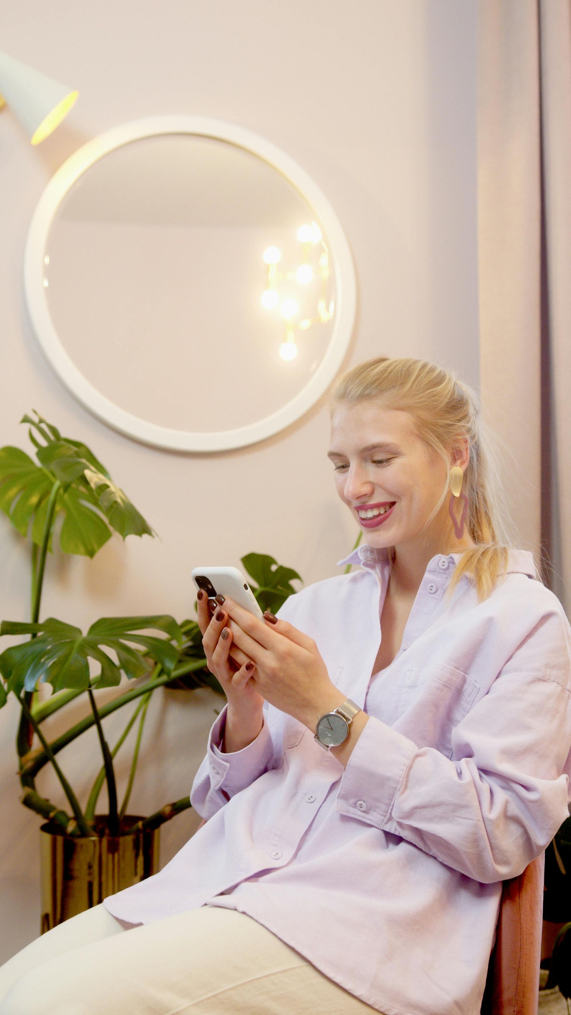 Une femme vêtue d'un blazer lilas clair sourit à son téléphone dans une pièce moderne