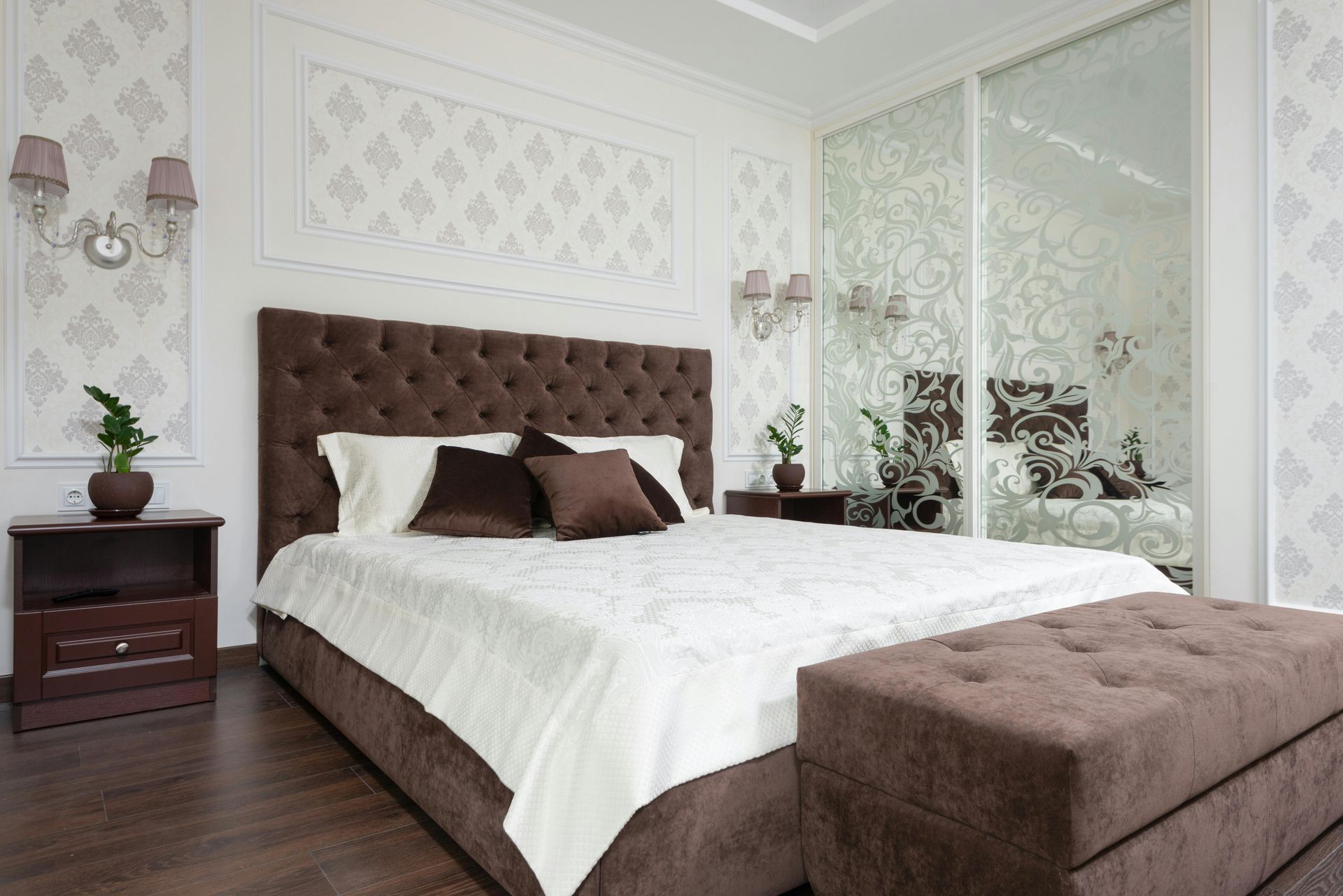 Chambre élégante avec un lit capitonné marron, du linge de lit blanc, des panneaux muraux en miroir et un banc assorti.