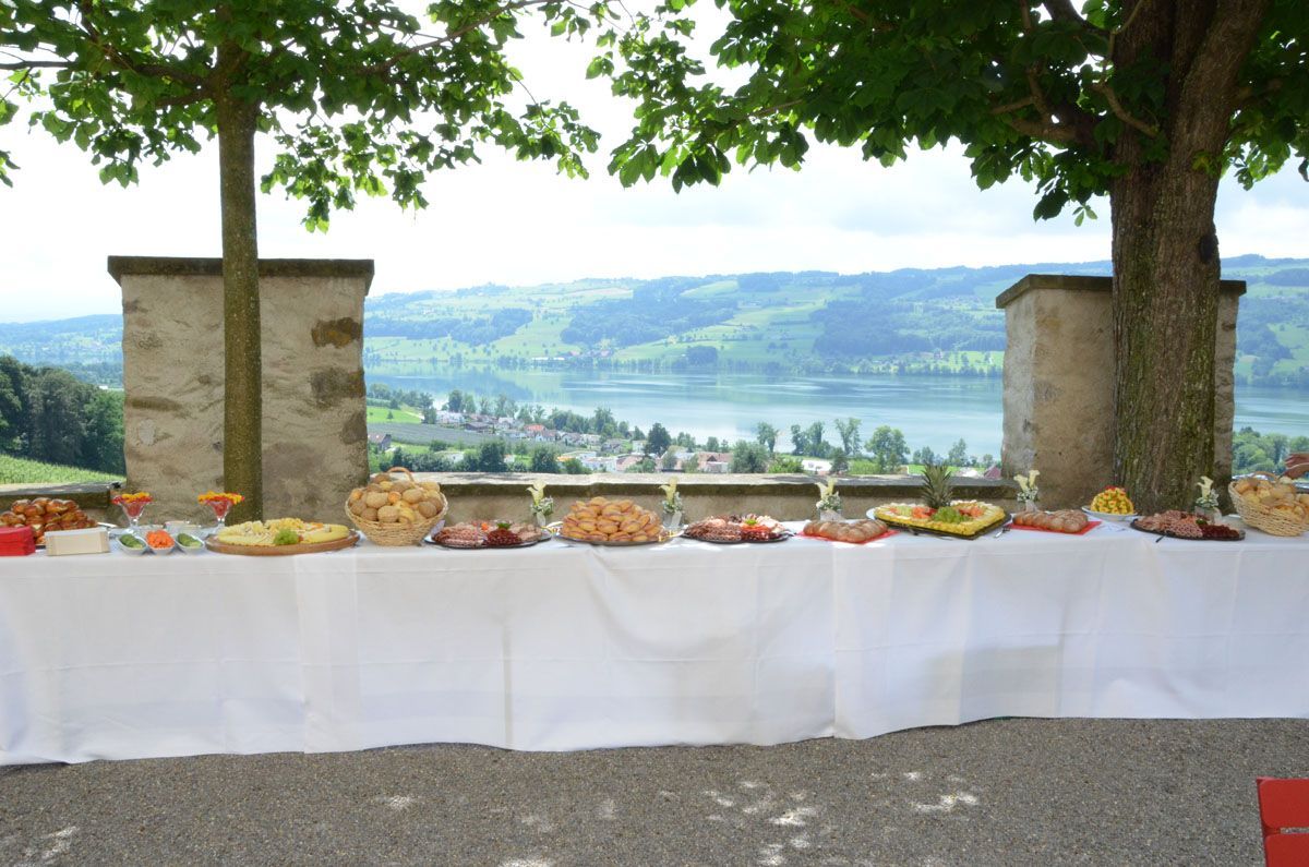 Langer Buffetisch mit weißer Tischdecke und Obst, unter Bäumen mit Blick auf ein malerisches Tal