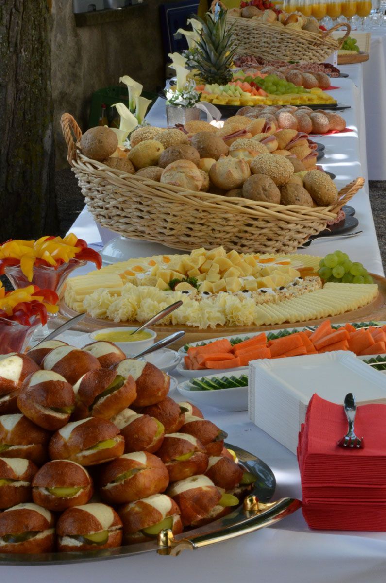 Buffet mit Körben voller Gebäck, Brötchen, Sandwiches und Obst, auf einer weißen Tischdecke angerichtet.