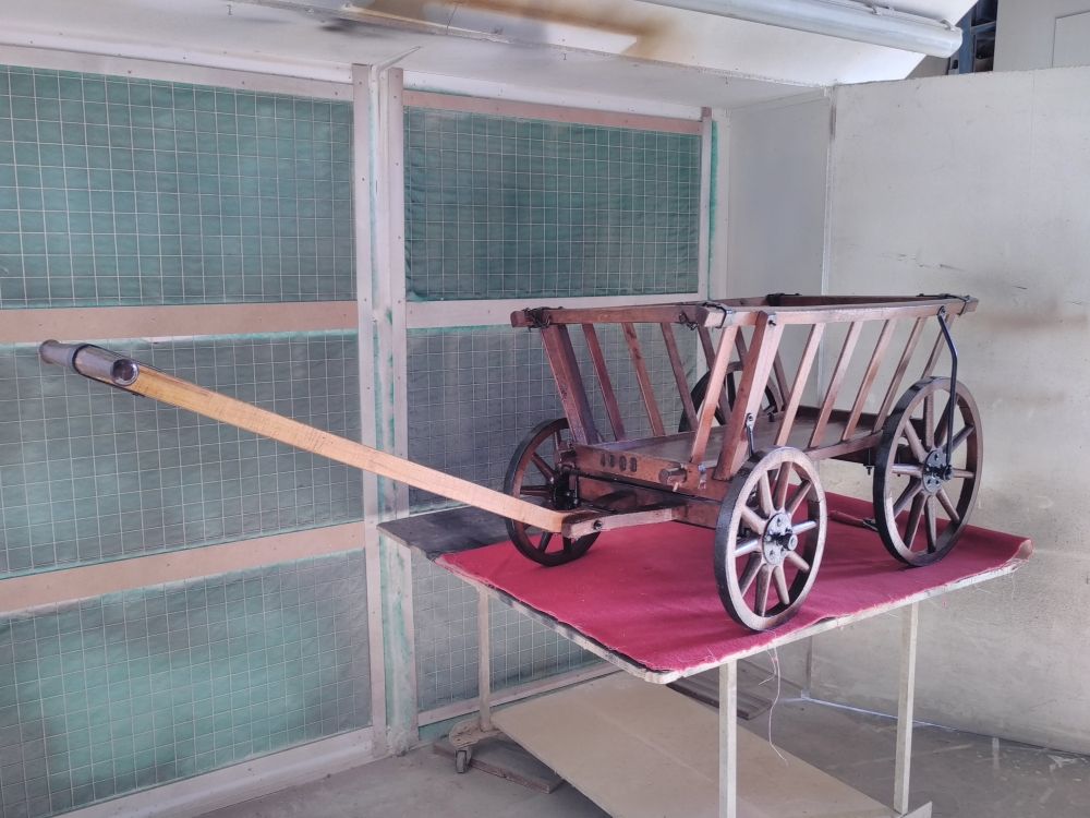 Dans un atelier, une maquette de chariot en bois à quatre roues et à long manche.