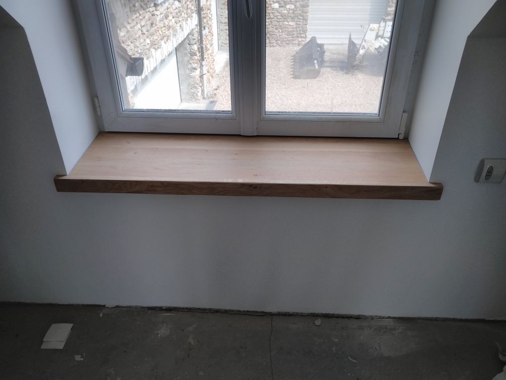 Un rebord de fenêtre en bois clair installé contre un mur blanc sous un cadre de fenêtre, au-dessus d'un sol en béton brut.