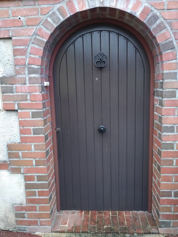 Une porte en bois sombre et cintrée, avec des planches verticales, un heurtoir noir et une poignée centrale, encastrée dans une arche en briques.